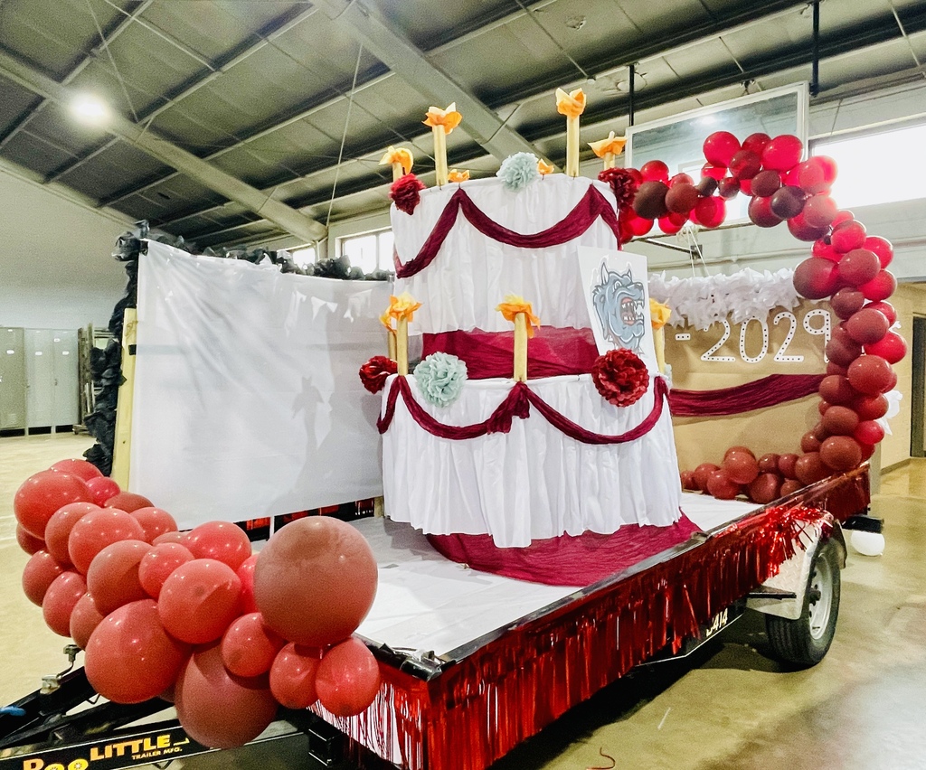 A red and white float on a trailer. The float is shaped like a cake. It has balloons and a banner that says 2029.