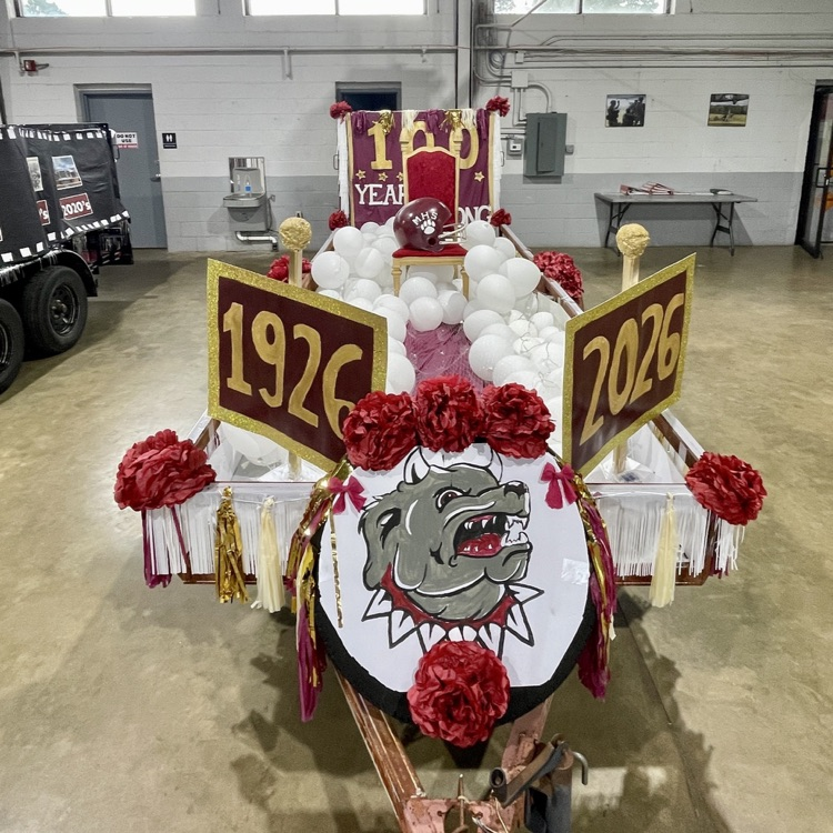 Decorated float with a dog mascot, red flowers, and white balloons. It has signs for years 1926 and 2026.