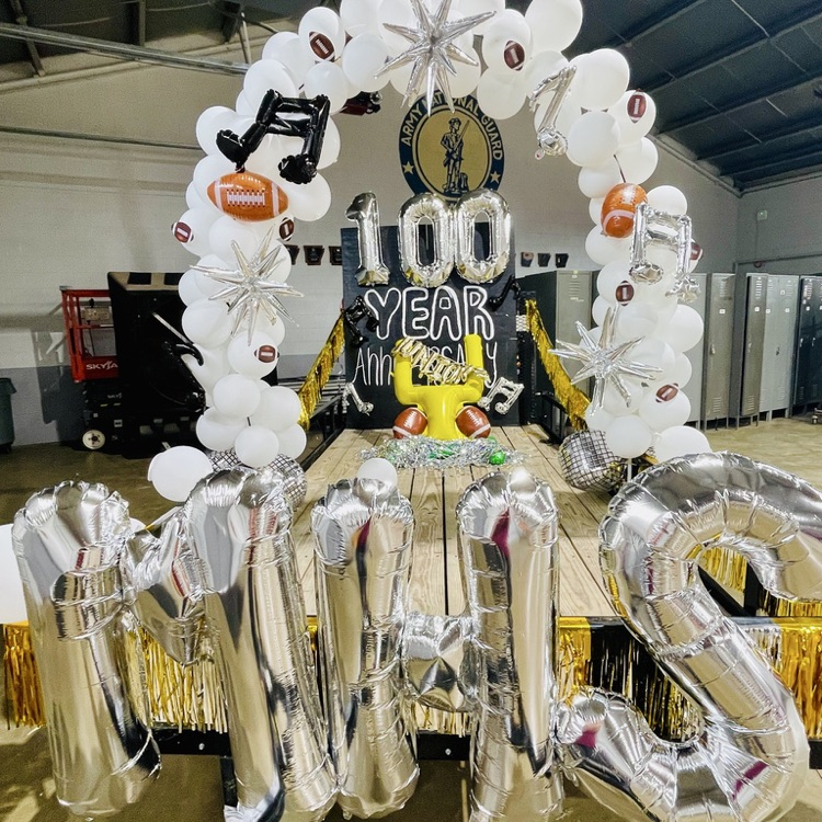 A room with a large white and silver balloon arch reading "100 Year". The arch has footballs and stars.