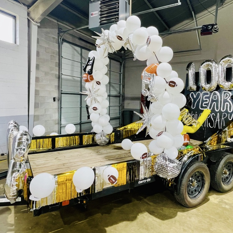 A trailer displays a large arch of white balloons, along with a banner reading "100 Year". The trailer is situated inside a building.