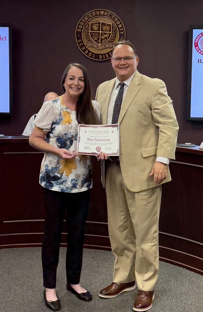 A man and woman stand in an office, presenting an award. The woman holds a certificate, smiling. Behind them, a wooden counter and monitors are visible.
