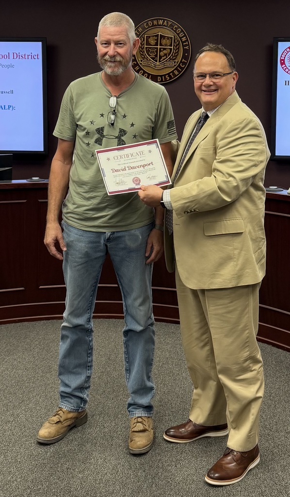 Two men standing in a room, one holding a certificate, wearing light-colored suits and jeans, and smiling.