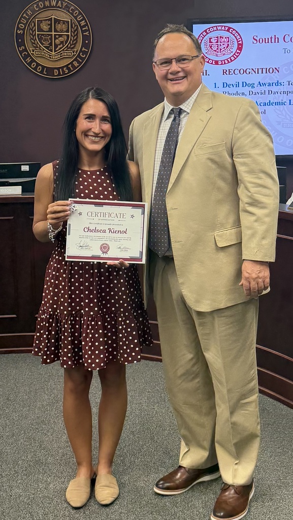 A woman holds a certificate while standing with a man, both smiling, in front of a desk.