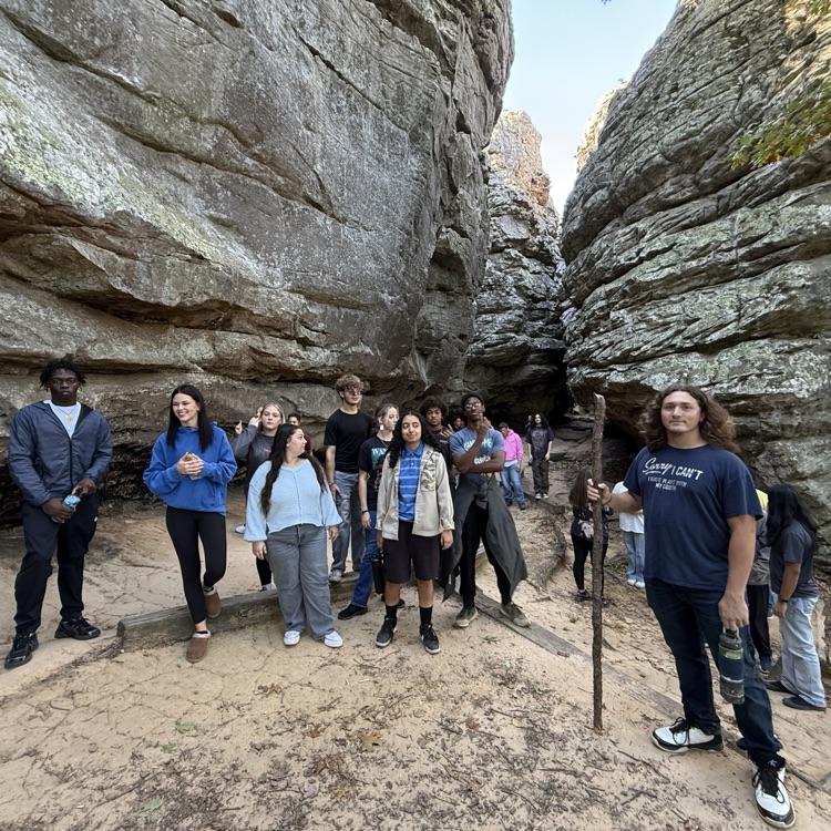 Students in casual attire standing in a rocky canyon, some holding a stick, with a natural background.