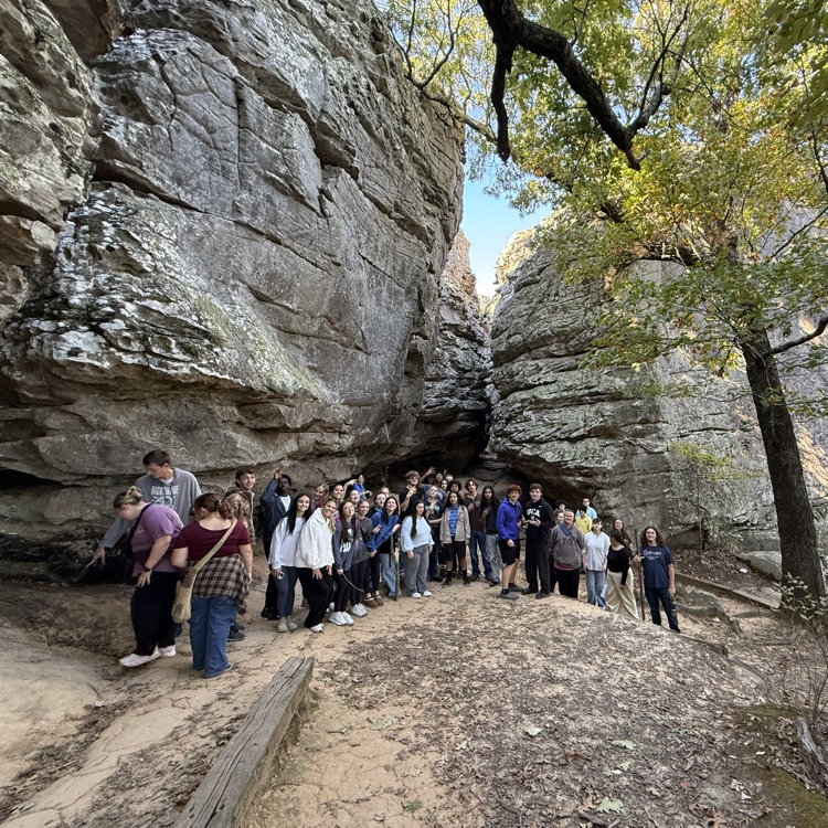 A group of people in front of a rocky cliff with a tree, some wearing hats.