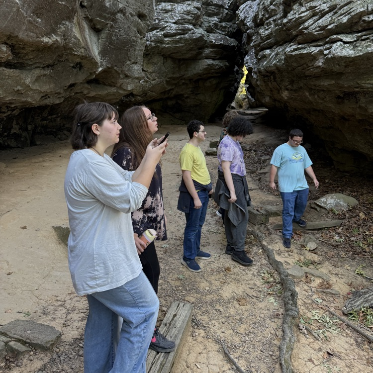 Five people on a dirt path. One person holds a phone. Rocks and rock walls in background.