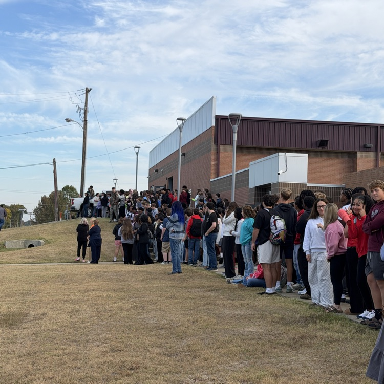 People stand in line on a dry grassy field with a school building in the background.