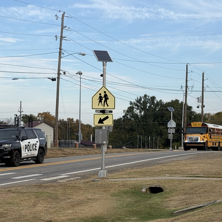 Police SUV parked on road, pedestrian crossing sign, school bus driving, man standing near road, utility poles and wires.