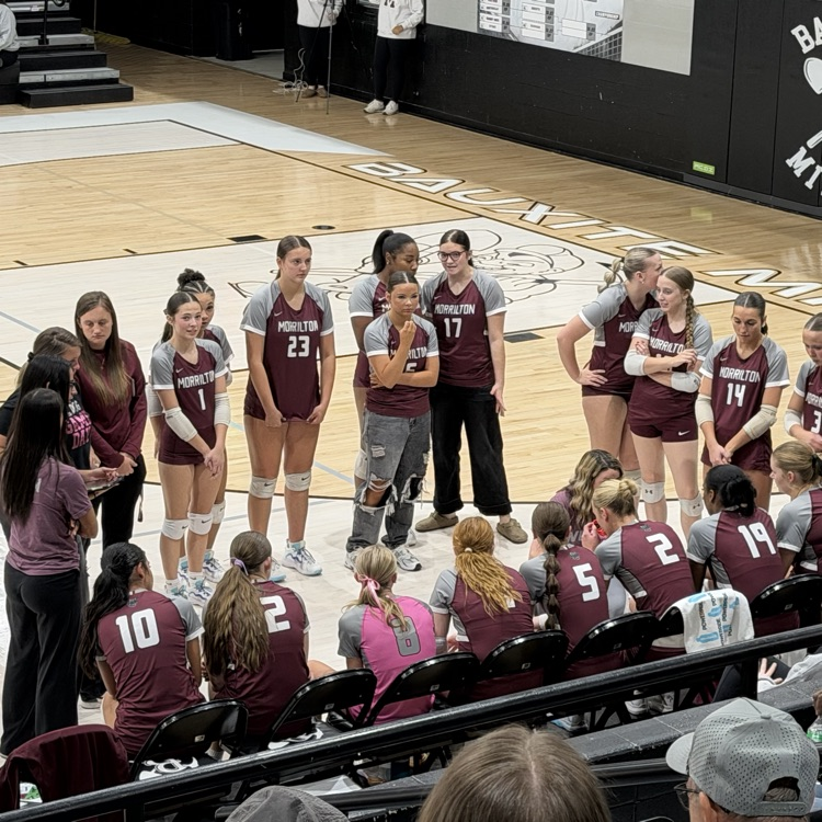 Volleyball team gathers around coach on court with audience in bleachers, wearing maroon uniforms.