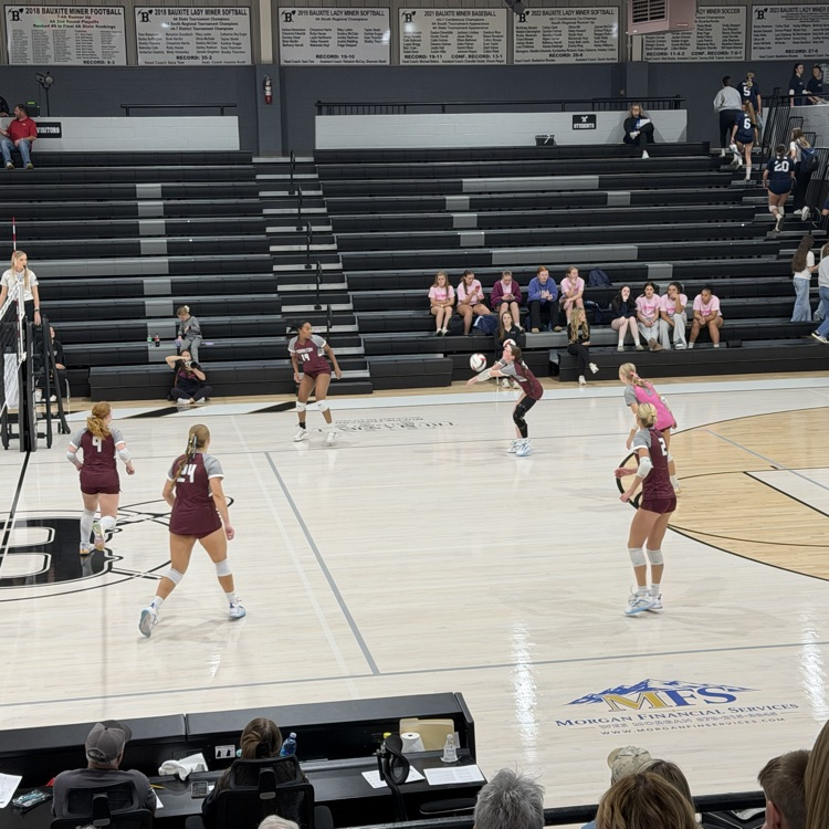 A group of people are playing volleyball on an indoor court. Spectators sit on the bleachers.