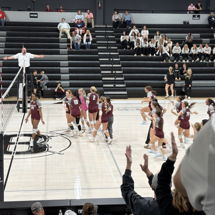 A group of young girls playing volleyball with a referee. Spectators sit on bleachers.