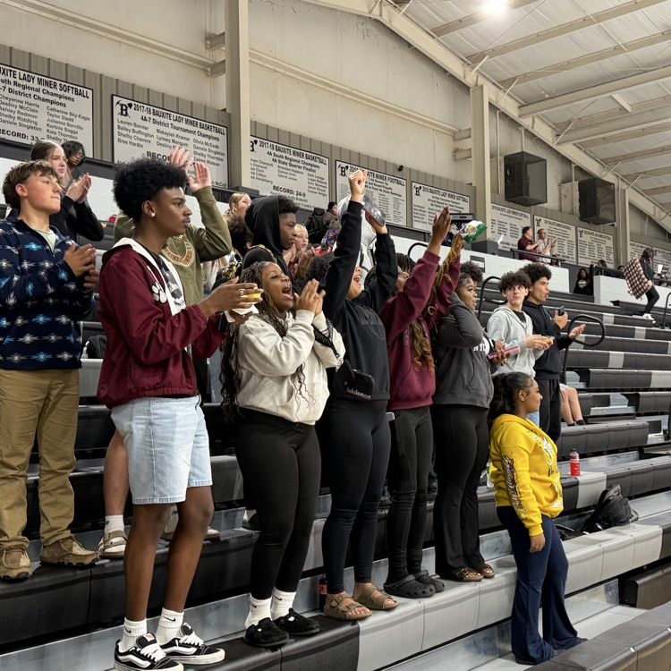 Students in a gym, some clapping and others standing on bleachers, some with hands raised.