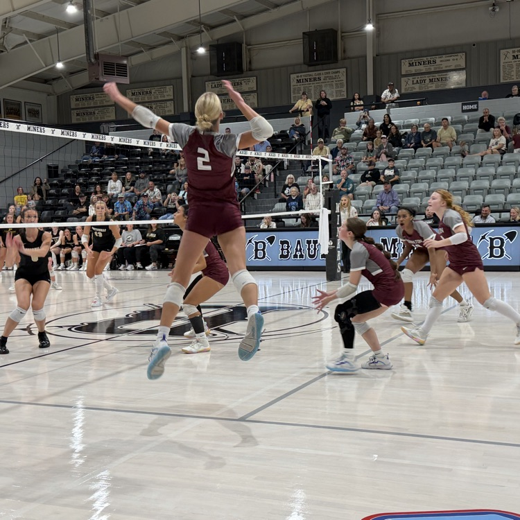 Volleyball players in maroon uniforms jump and play indoors on a court. A player with number 2 leaps to block a ball.