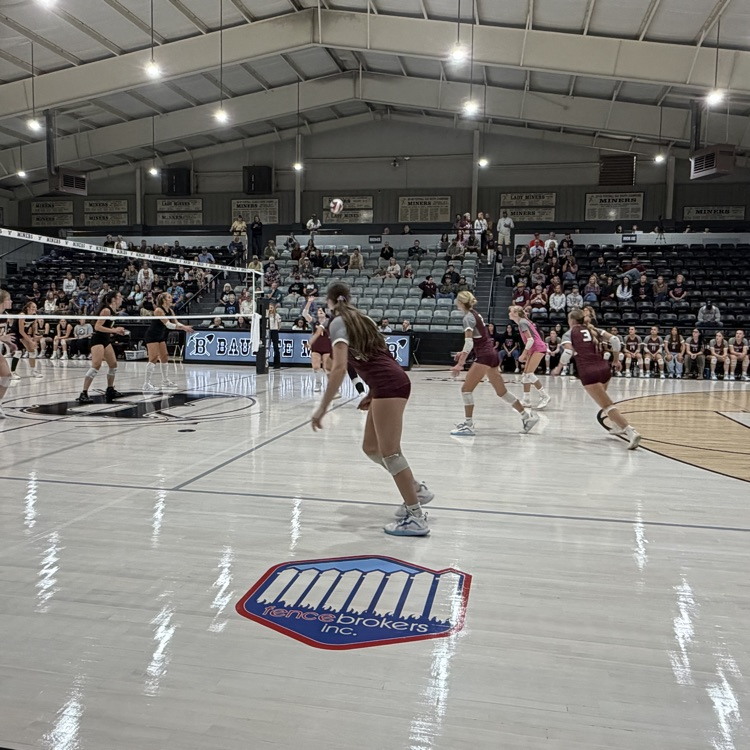 A group of girls in maroon uniforms play volleyball on a court with a logo. Spectators watch from the stands.