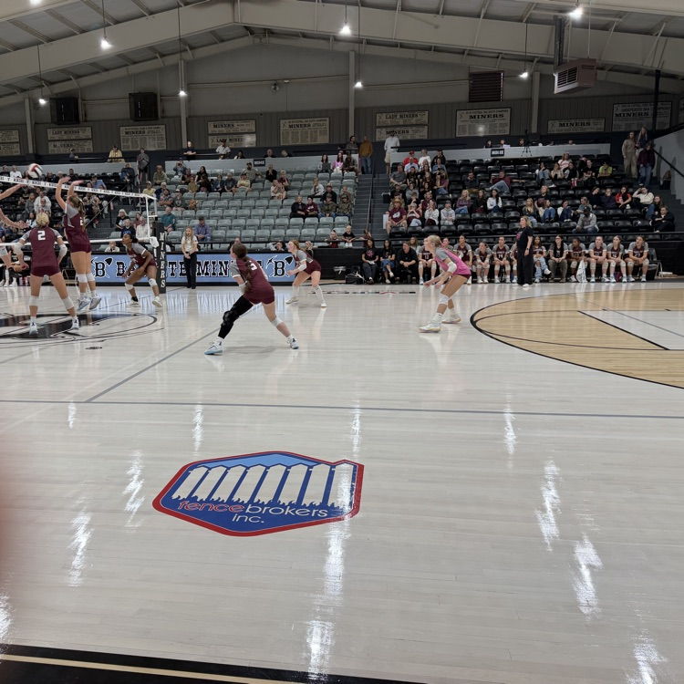 People playing volleyball on a court with one person jumping. People seated on bleachers.