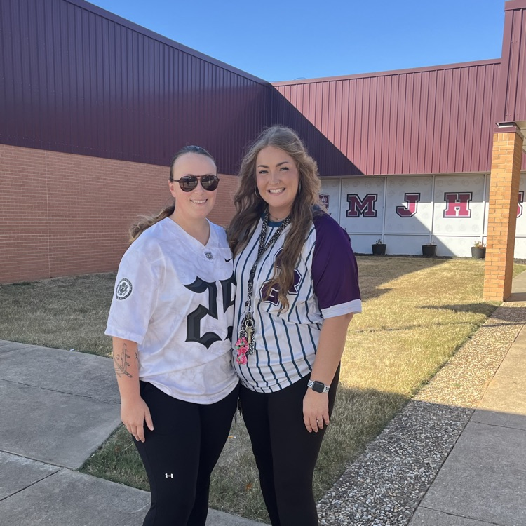 Two women stand side by side in athletic wear and sneakers. One wears a white jersey with the number 21. Behind them is a building with a flagpole and brick pillar.