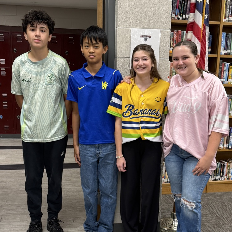 Four students stand in a row in a library, with one holding a baseball jersey.