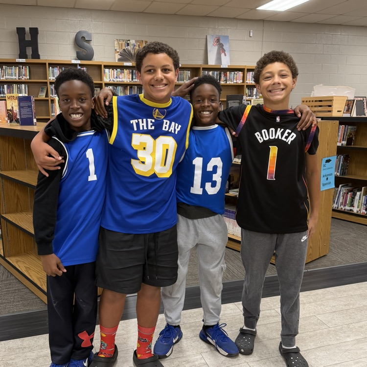 Four boys in sports jerseys standing together in front of library shelves. One wearing blue, one orange, one black, and one white.