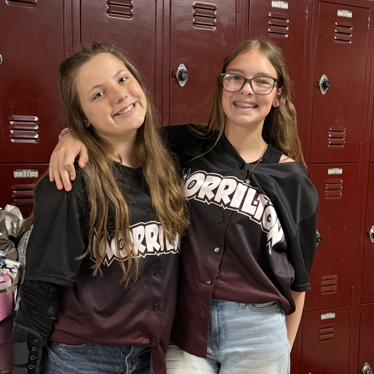 Two young girls, both wearing maroon and black baseball jerseys with the word "Morrilton" on them, pose for a picture in front of red lockers.