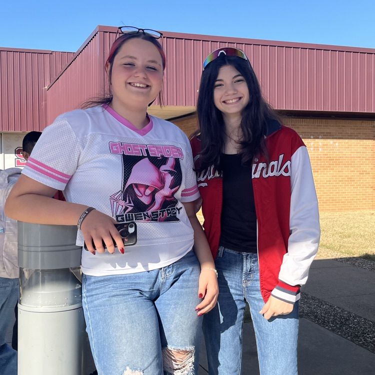 Two students stand by a pole outside a school building; one holds a phone, the other wears a red jacket.