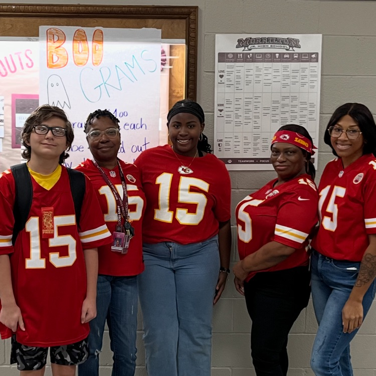 Five people wearing red jerseys with numbers standing in front of a wall with two signs.