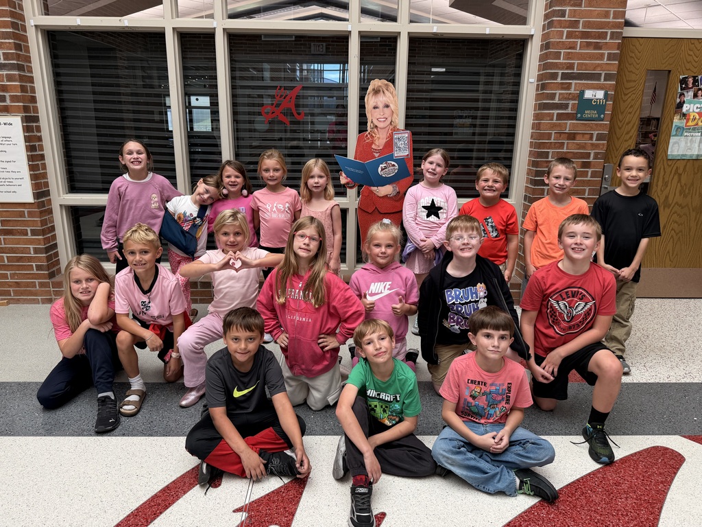 Aurora Elementary students in pink posing with a Dolly Parton Imagination Library cutout outside the school media center.