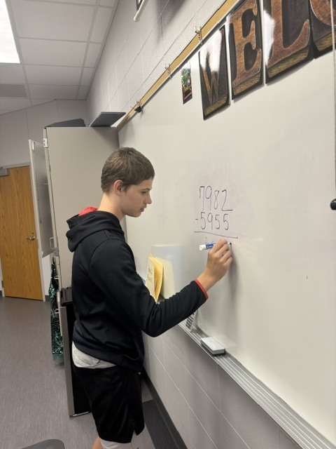 High school student in a black hoodie works through a subtraction problem on the whiteboard during algebra board work.