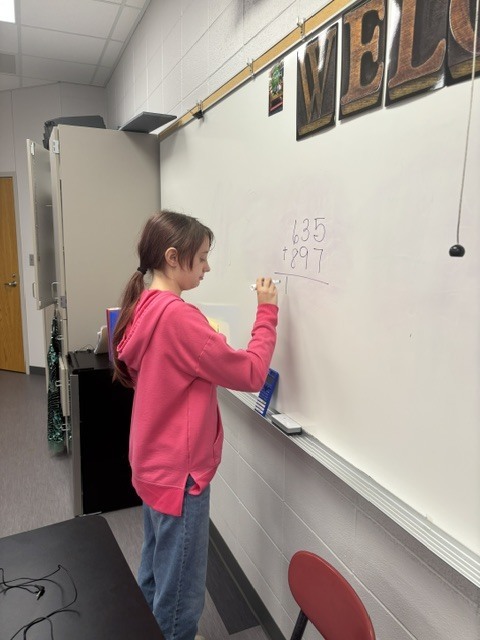 Student in a pink sweatshirt solves a multi-digit addition problem on the whiteboard in Mrs. Wuestefeld's Applied Algebra class.