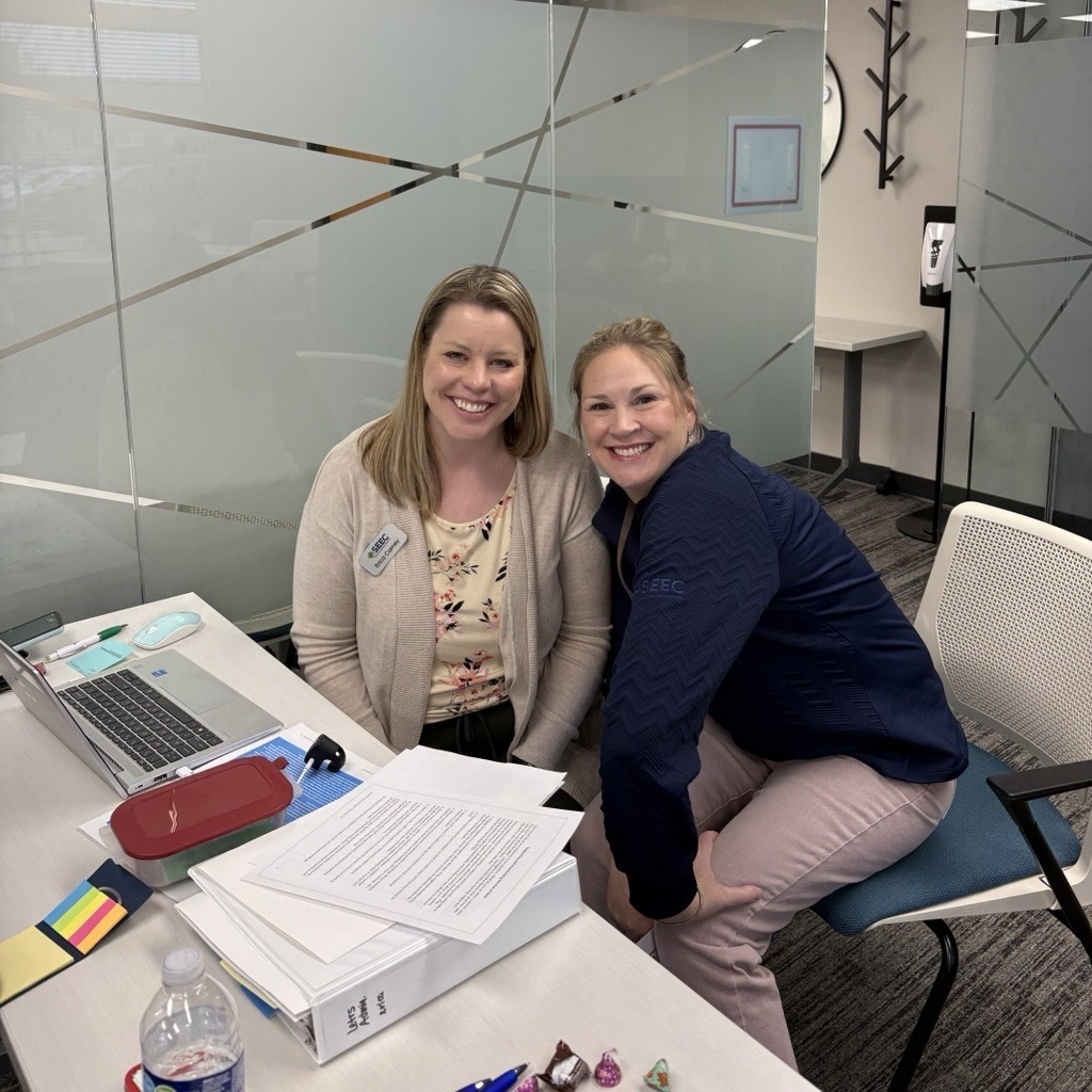 two trainers smiling and sitting at a table with paper and computer
