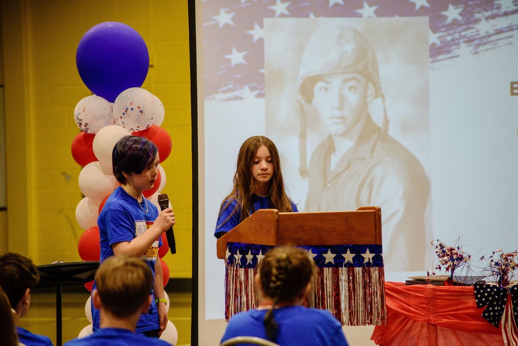 Two students giving a presentation at the Patriot Program Assembly as students in the audience listen.