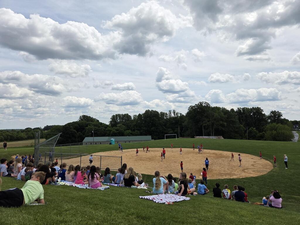 A large group of students sitting on a grassy hill watching staff and students play a kickball game.