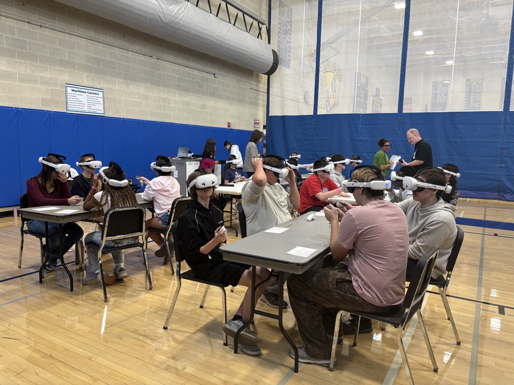 Students sitting at tables in a gymnasium. They are wearing virtual reality goggles and holding joysticks. They are participating in an activity related to STEM careers.
