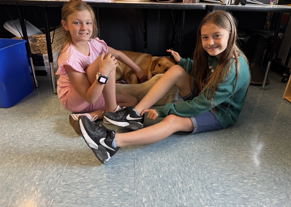 Two children sit on a tiled floor with a dog lying between them. The child on the left wears a pink dress and a smartwatch, while the child on the right wears a green sweatshirt and shorts. Their faces are obscured for privacy.