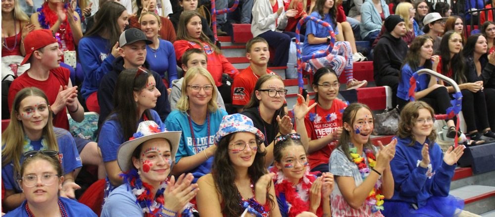 A diverse crowd of high school students in red, white, and blue spirit wear cheer from gymnasium bleachers.