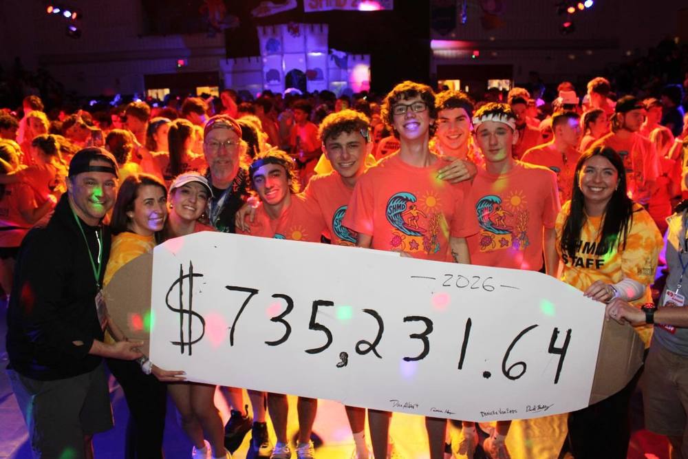 A large group of participants wearing matching event shirts stand together on a gym floor holding an oversized white sign displaying the total amount raised: $735,231.64 for 2026. Colorful stage lighting illuminates the crowded scene, and people surround the group in the background during the South High Marathon Dance.