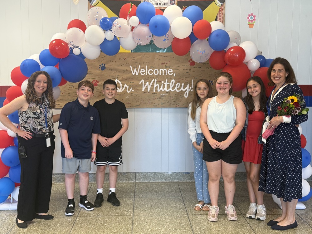A group of students and two women stand under a balloon arch, with a banner reading 'Welcome, Dr. Whitley!' behind them.