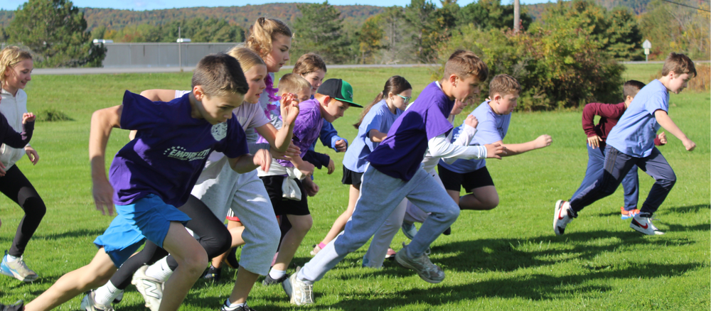 A group of children run energetically across a green grassy field on a sunny day.