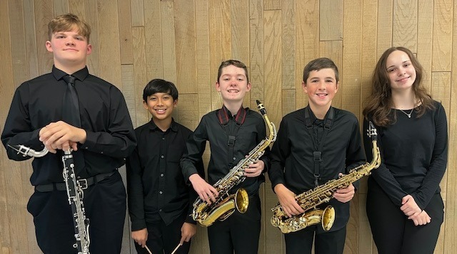 Five musicians dressed in black concert attire stand in front of a light wooden wall. From left to right: one person holds a bass clarinet, the next holds two mallets for percussion, followed by two individuals each holding an alto saxophone, and the last person stands without an instrument.