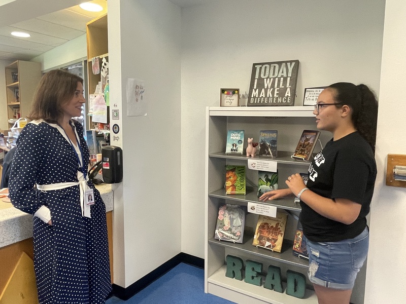 A woman in a polka dot dress talks to a young person standing by a bookshelf displaying children's books.