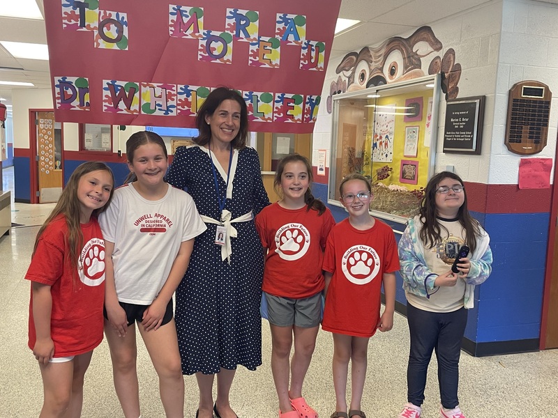 A woman in a polka dot dress smiles with five young girls in red and white t-shirts in a school hallway.