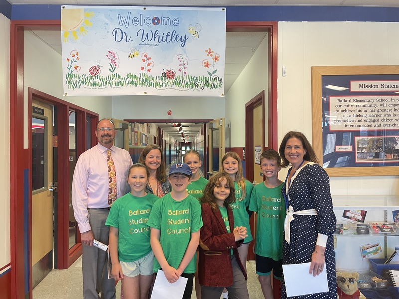 A group of students and two adults stand in a school hallway, posing for a photo in front of a "Welcome Dr. Whitley" banner.