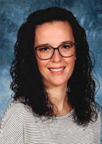 A woman with dark curly hair and glasses smiles warmly.