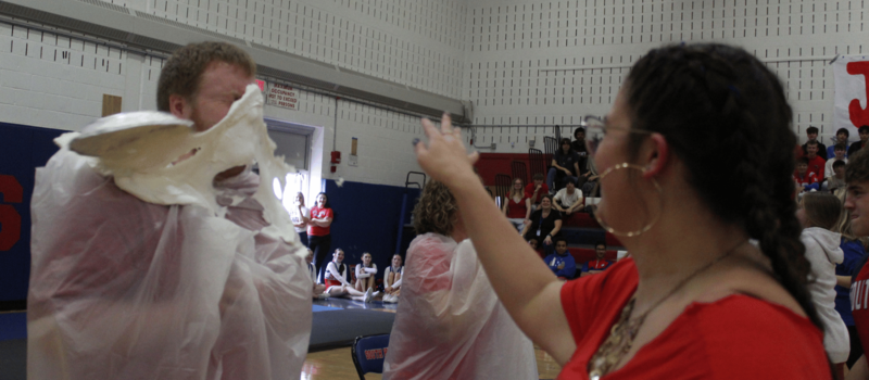 A person wearing a red shirt throws a pie into the face of someone wearing a plastic poncho in a gymnasium.