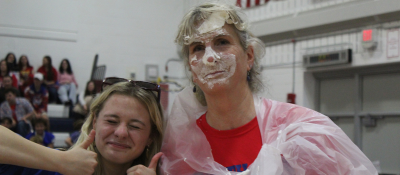 A smiling woman with whipped cream on her face poses indoors next to a younger woman giving two thumbs up.