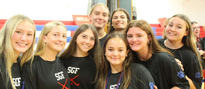 A group of smiling young women wearing matching black shirts with "SGF" and crossed sticks graphics pose together indoors.