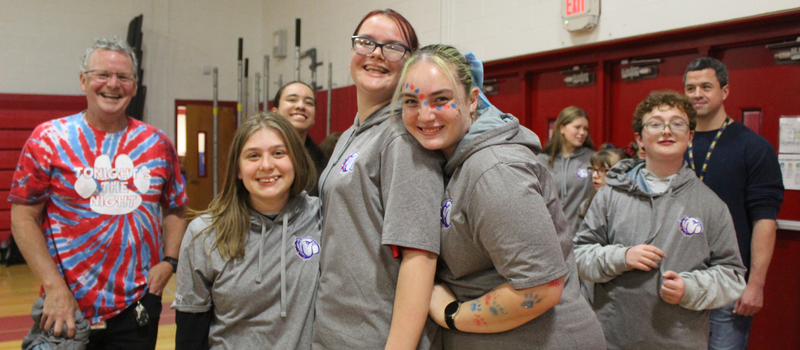A group of students and teachers in grey hoodies and tie-dye shirts smile together inside a school gym.