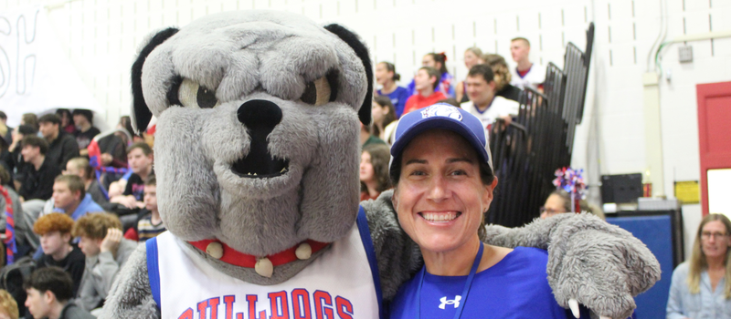 A smiling woman in a blue baseball cap poses with a gray bulldog mascot in a crowded gymnasium.