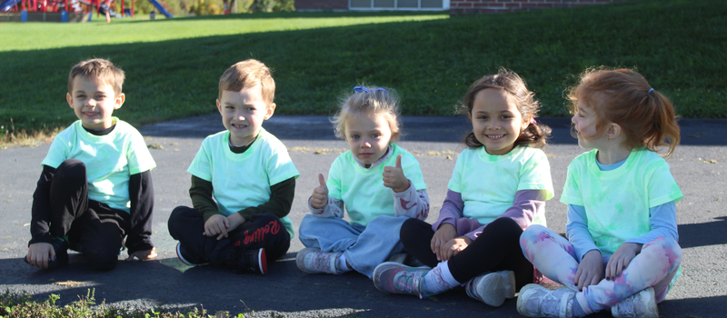 Five young children in matching light green shirts sit cross-legged outdoors.