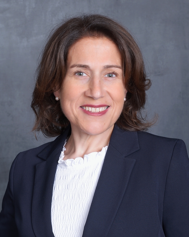 A woman with brown hair smiles warmly, wearing a dark blazer over a white ruffled shirt.