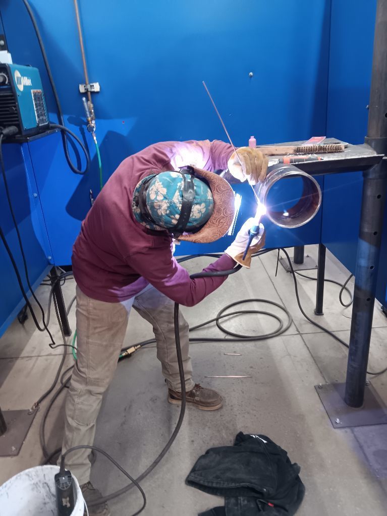 Students participating in a welding competition, wearing protective welding helmets and gear while working on pipe welding projects in a shop environment. 1